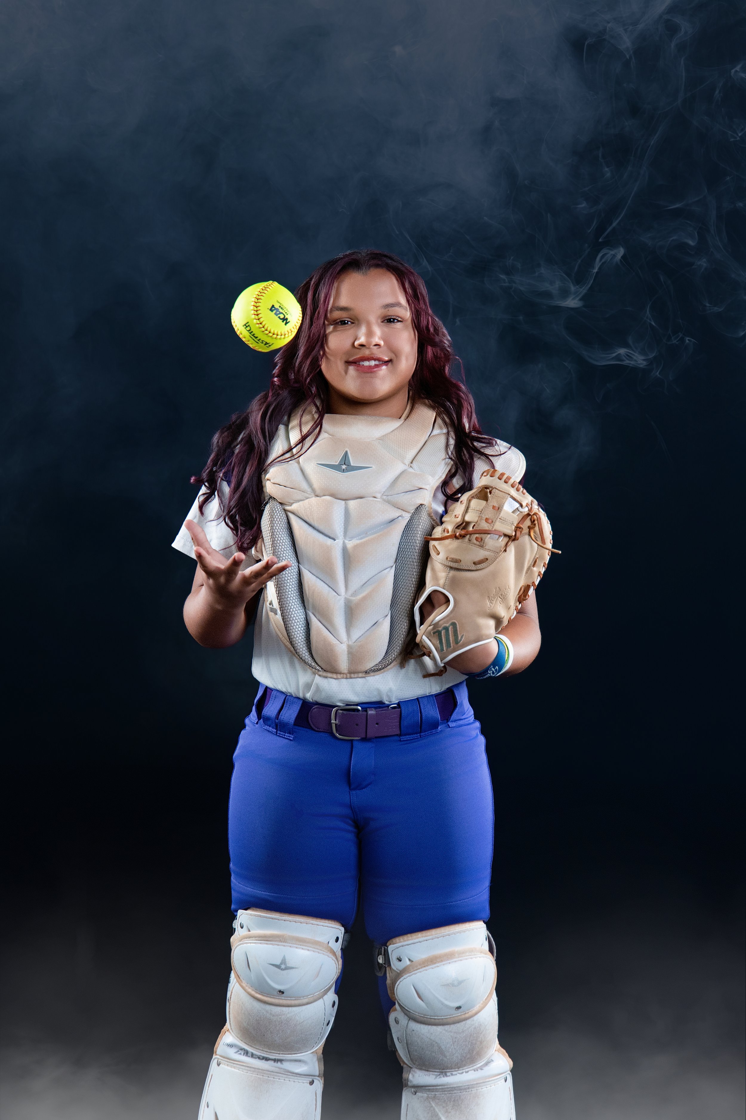 Young female softball catcher in uniform, wearing catching gear, holding a baseball glove, with a softball in midair behind her, against a dark background.