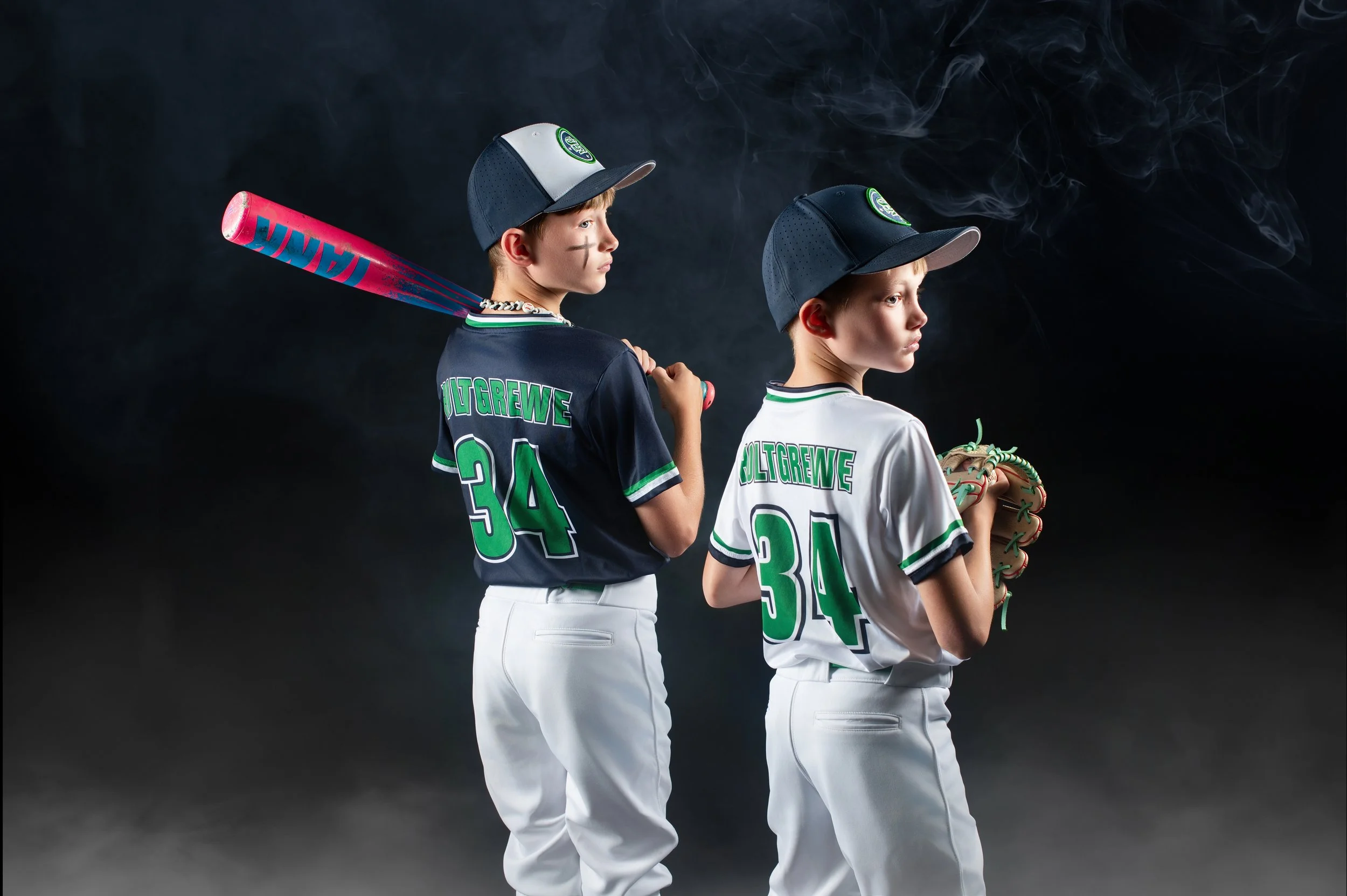 Two young boys in sports uniforms posing with one holding a baseball bat and the other holding a baseball glove. Both wear caps and jerseys with green and white accents, against a dark background with smoke.