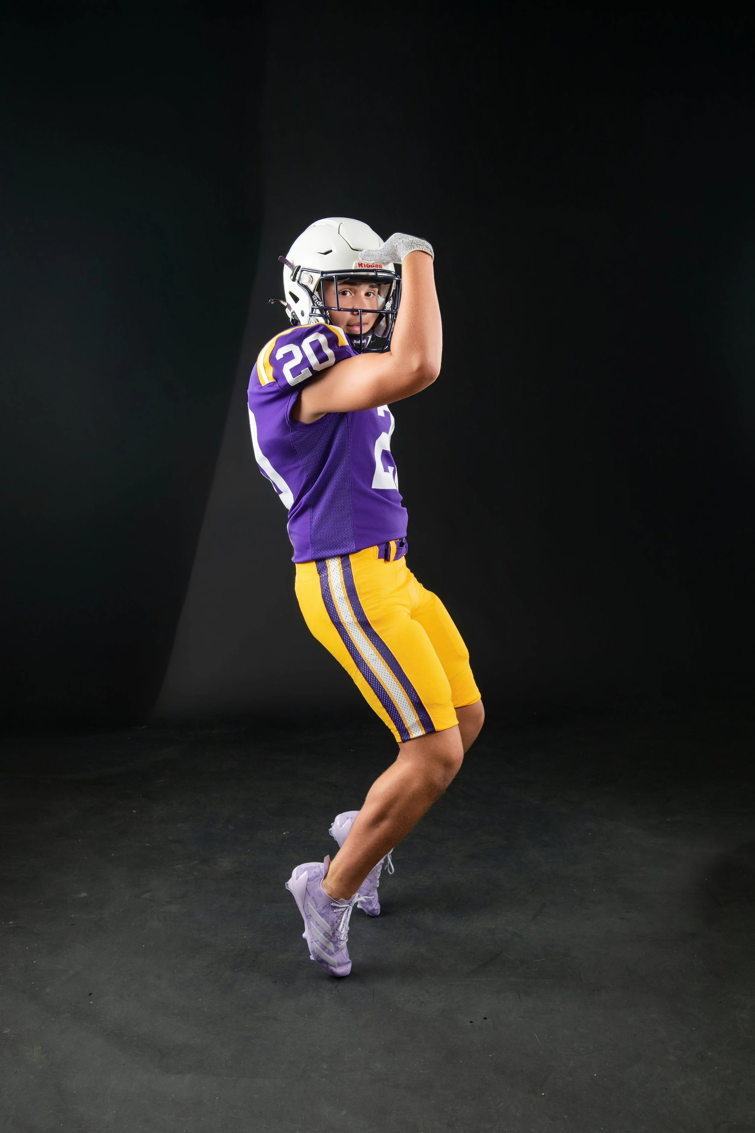 A young male athlete wearing a purple jersey with the number 20, yellow athletic shorts with purple stripes, a white football helmet, and purple sneakers, standing on a black background.