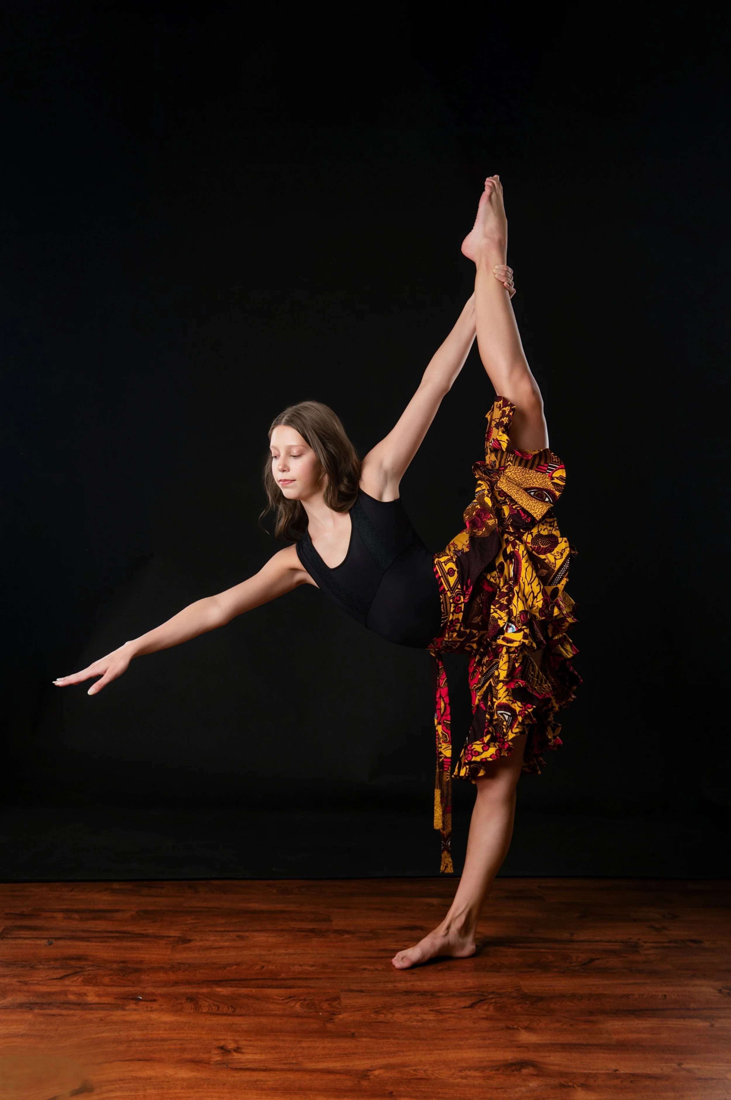 A woman in a black leotard and colorful wrap skirt practicing a ballet pose, standing on one leg with the other leg extended upward and holding it with her hand against a black backdrop, on a wooden floor.