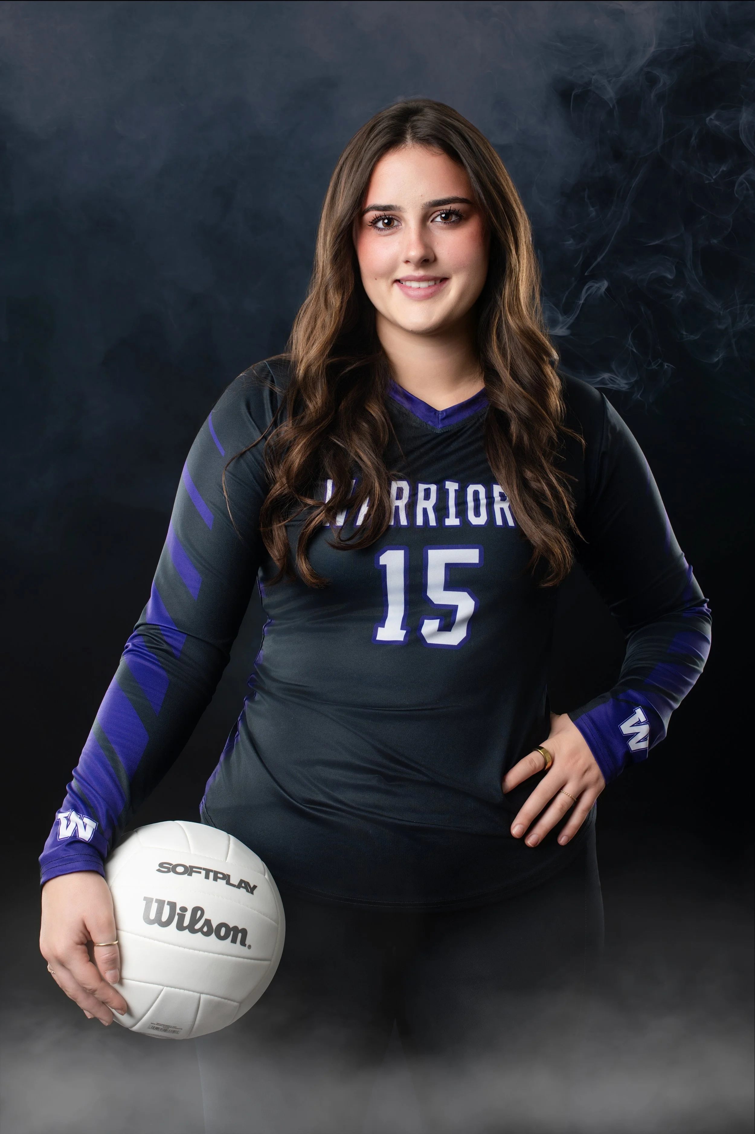 A young woman in a blue and black volleyball uniform with the number 15, holding a white Wilson volleyball, standing against a dark smoky background.
