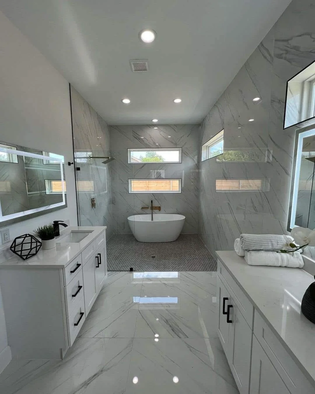 Modern bathroom with white marble walls and floors, a free-standing bathtub, two horizontal windows, under-cabinet lighting, a vanity with a sink, black hardware, and neatly folded towels.