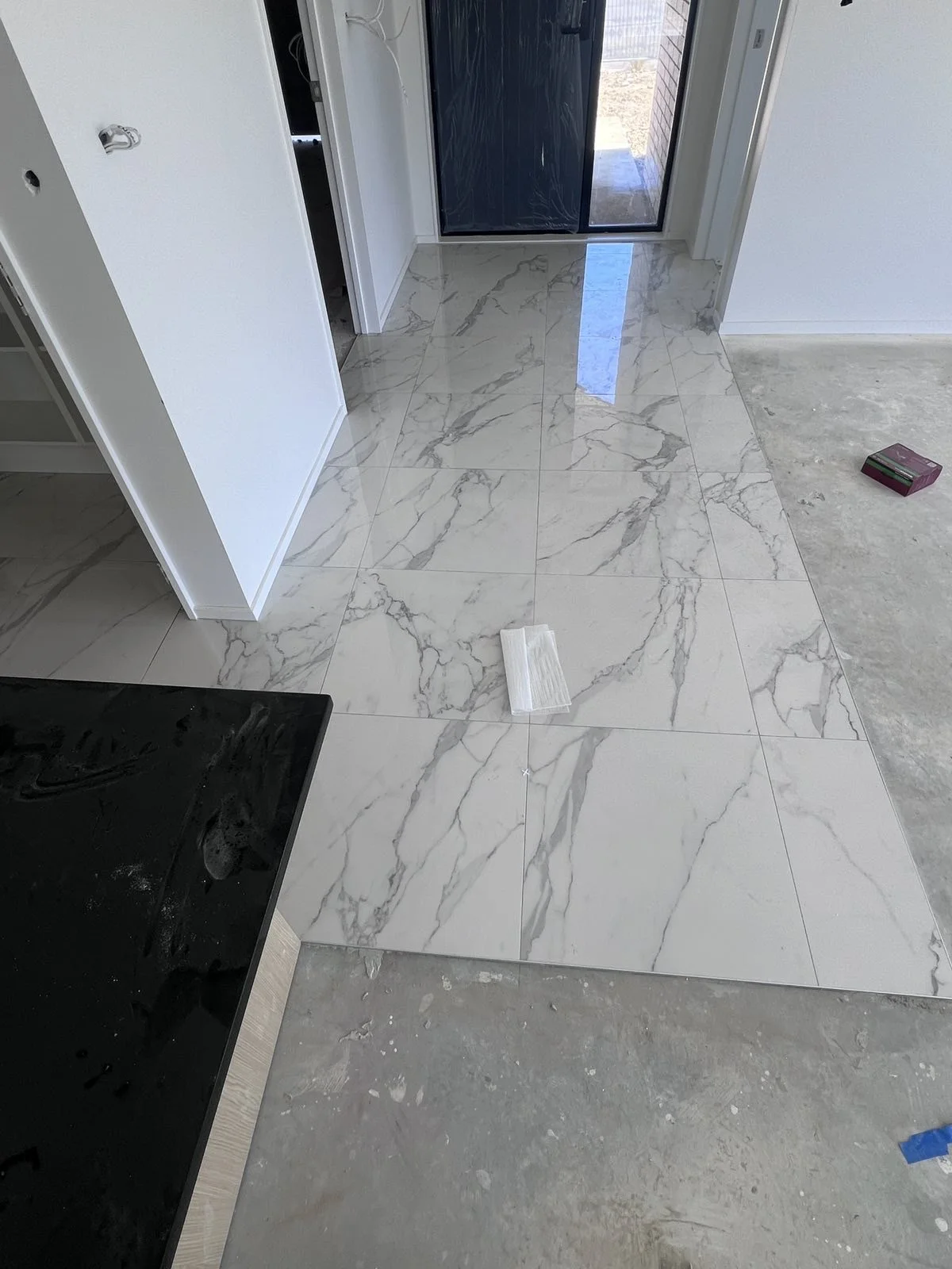 Interior of a room with newly installed marble tile flooring near a door, with some construction materials and tools visible, including a black countertop, a white sponge, and a box on the floor.
