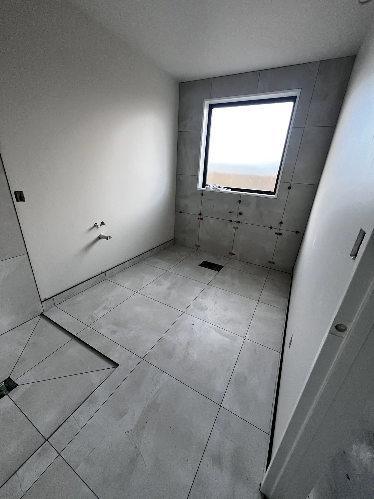 An unfinished bathroom with white tiled walls and floor, a window, and plumbing fixtures for a toilet and sink.