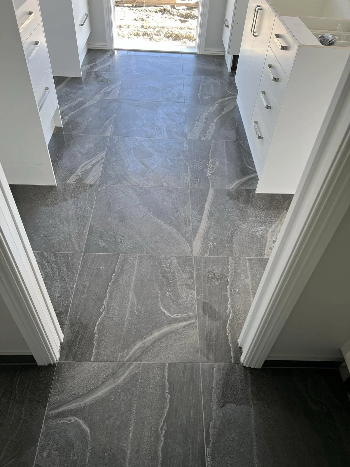A view of gray marble tile flooring in a modern kitchen with white cabinets and a sliding glass door leading outside.