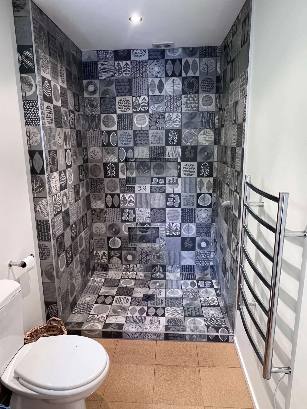 Bathroom with a walk-in shower featuring walls and floor covered in black, white, and gray patterned tiles, a white toilet, a woven basket near the toilet, and a chrome towel rack on the wall.