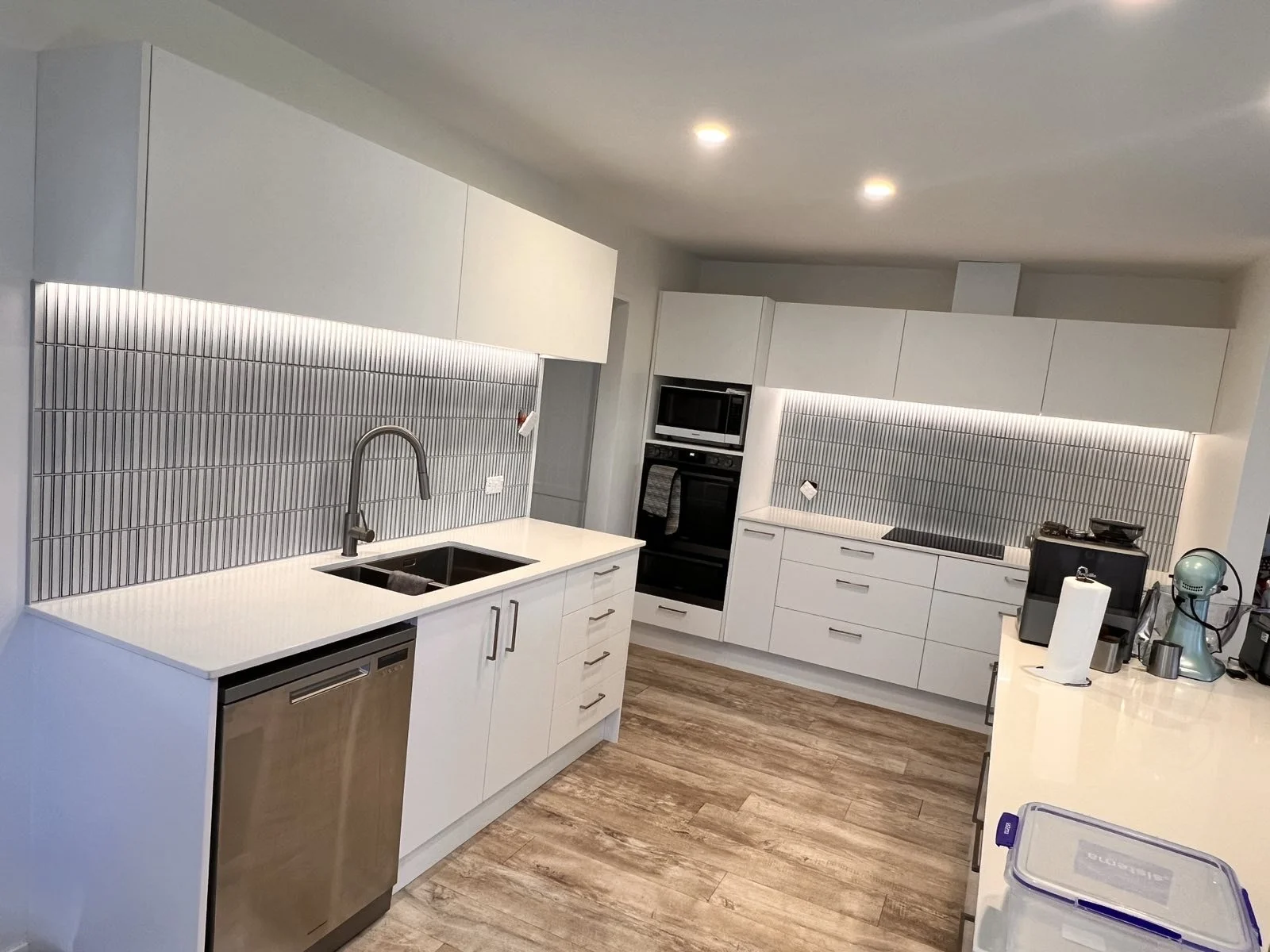 Modern kitchen with white cabinets, gray tile backsplash, and wood-look flooring, featuring a sink, dishwasher, oven, microwave, and various countertop appliances.