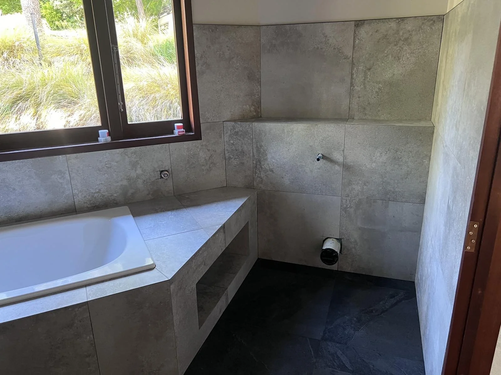 A bathroom under construction with tile walls, a corner bathtub, an open window with a view of greenery, and visible plumbing fixtures.