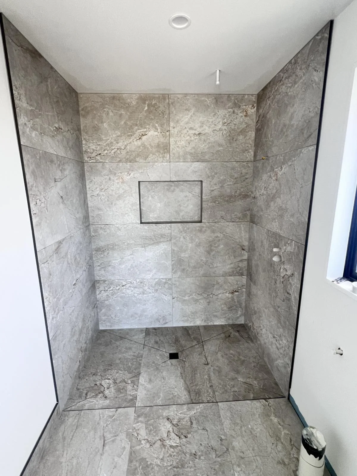 A partially finished shower with gray marble tiles on the walls and floor, a small recessed square shelf in the wall, a drain on the floor, and an opening for plumbing fixtures. The edges are outlined with black trim.