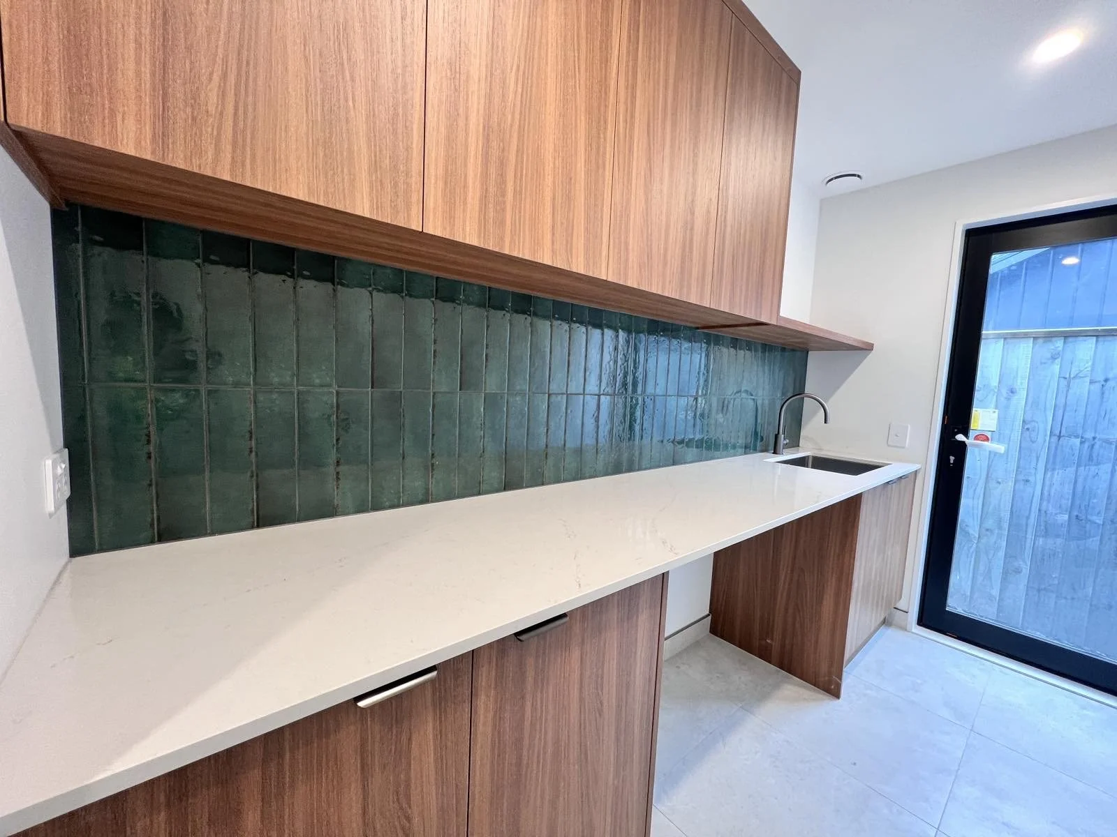 Modern kitchen with white countertop, wooden cabinets, green tiled backsplash, and a black sink with a chrome faucet. A glass door leads outside.