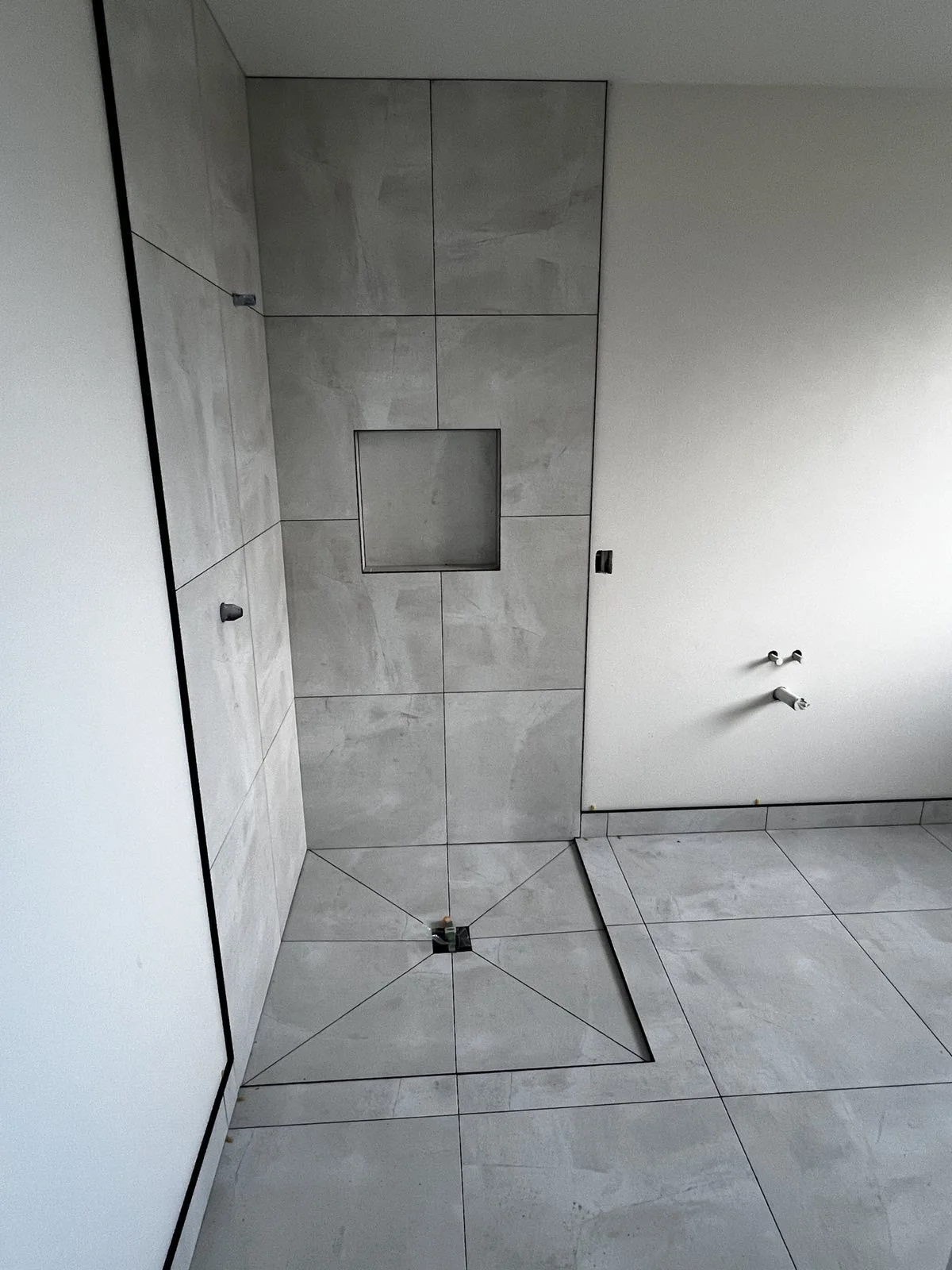 A bathroom under construction with tiled walls and floors, a square shower niche in the tiled wall, and plumbing fixtures without sinks or faucets installed.