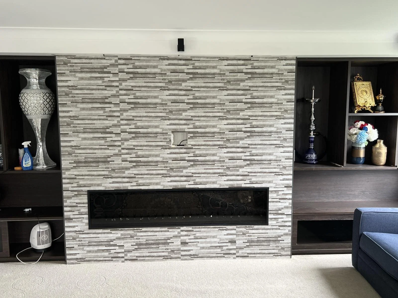 Living room with a modern gray stone fireplace, black shelving units on both sides displaying decorative items, a blue armchair partially visible on the right, and a beige carpeted floor.