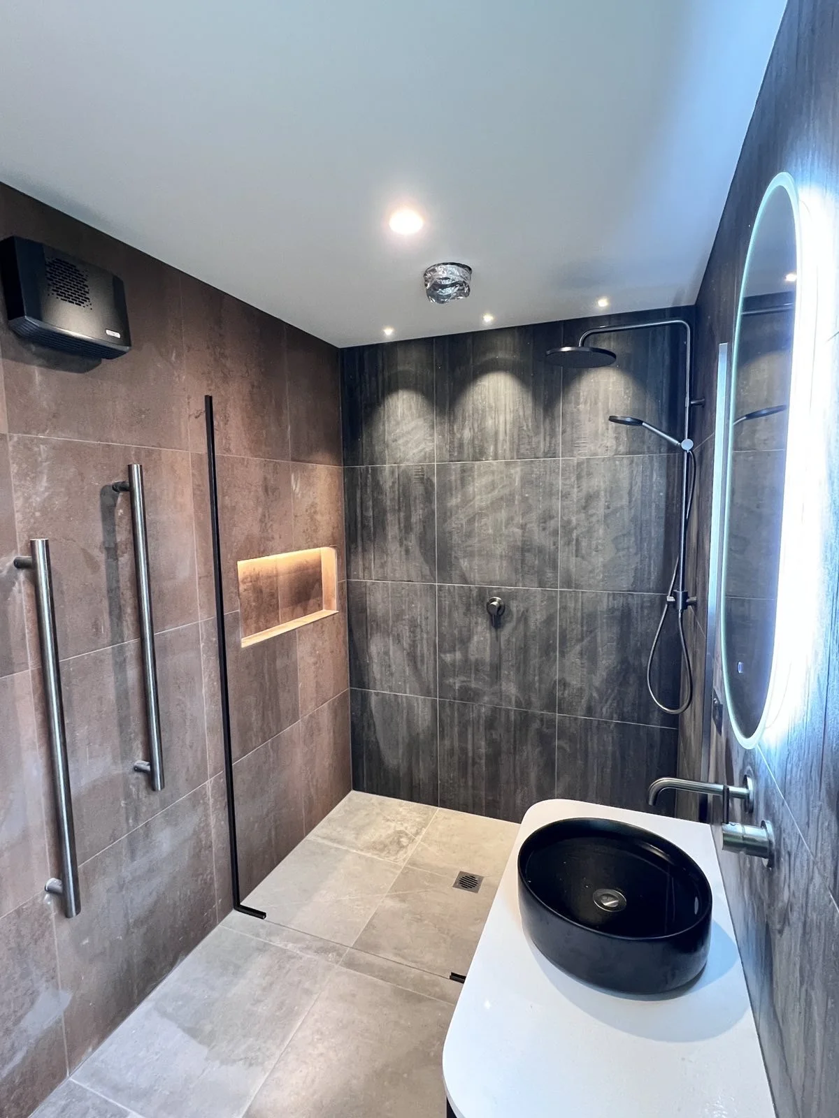 Modern bathroom with a walk-in shower featuring gray tiles, a black rain shower head and handheld shower, a small illuminated niche, and a black vessel sink on a white countertop next to a mirror with LED lighting.