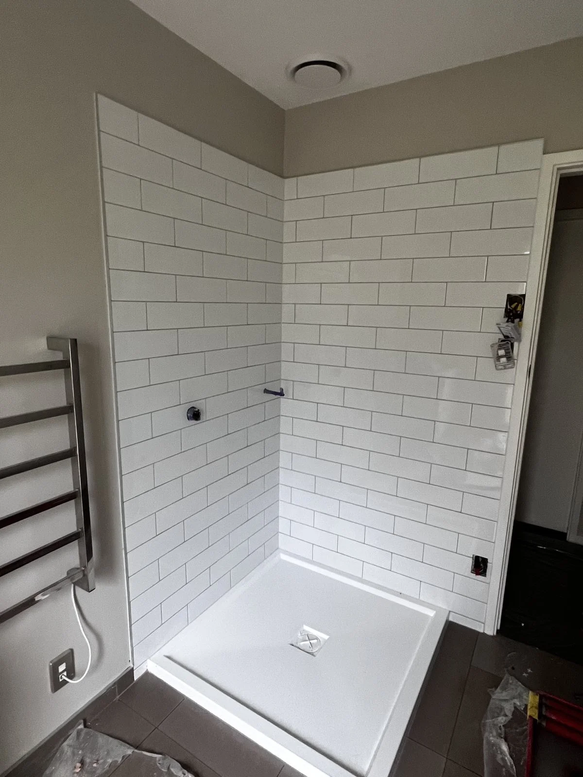 An unfinished bathroom shower area with white subway tile walls, a white shower base, and a dark tile floor. Electrical outlets and wall fixtures are not yet installed.