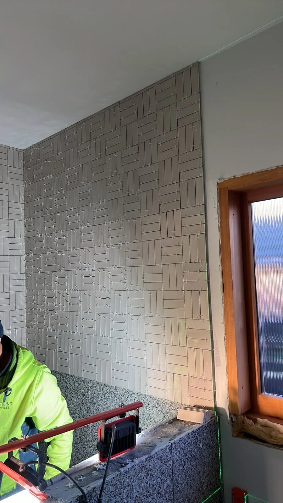 Interior view of a construction site with tiled wall, wooden window frame, and construction worker in a neon green jacket.