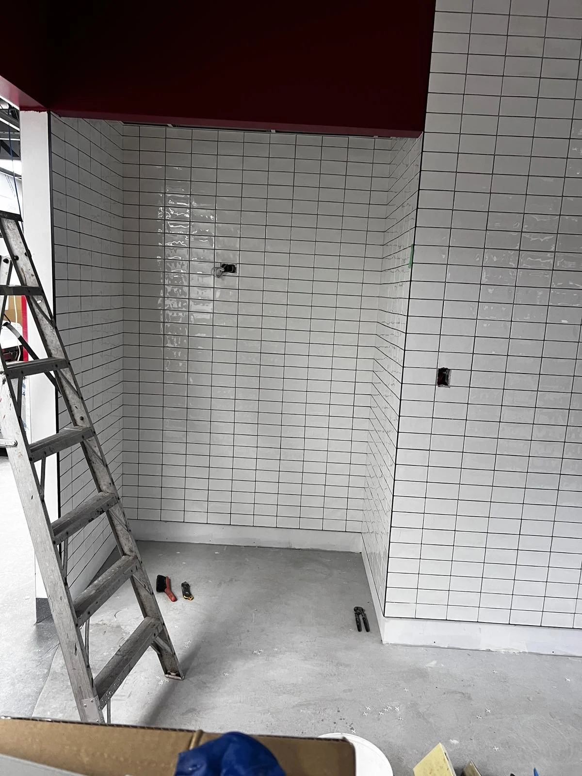 Interior space under construction with white tiled walls, electrical outlets, and tools on the floor, including a ladder leaning against the wall.