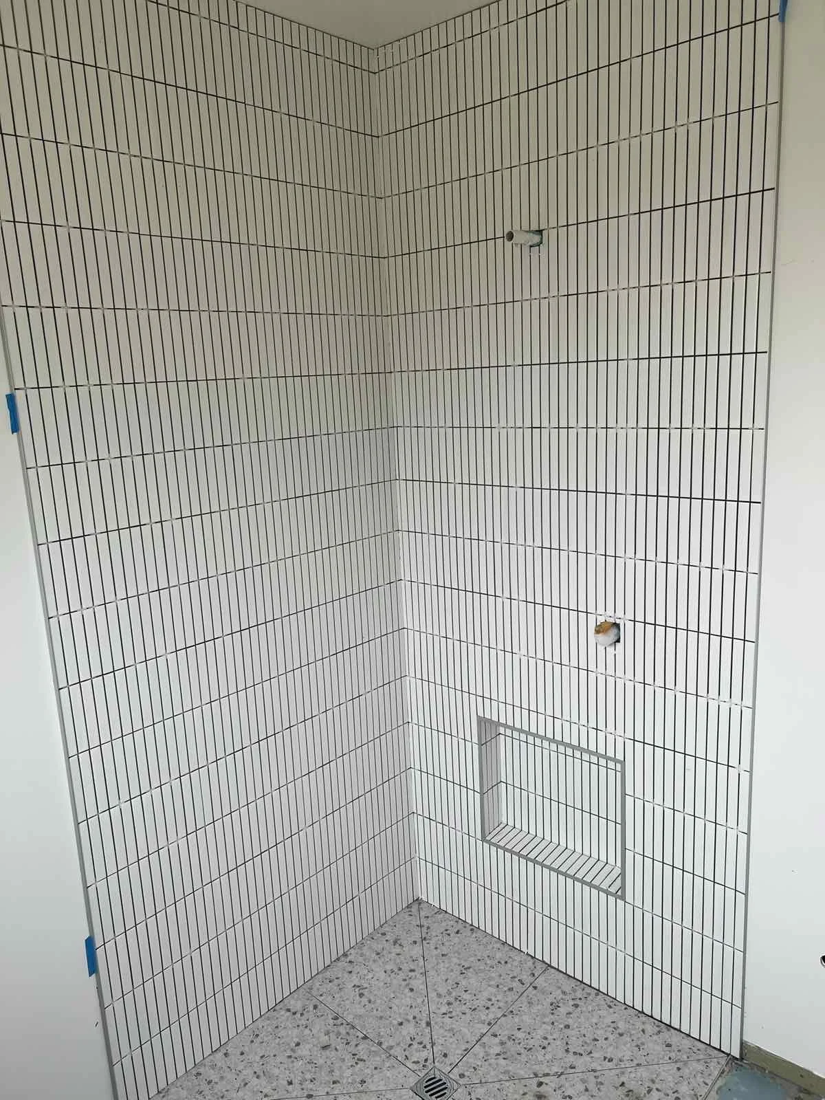 A corner of a bathroom shower with white rectangular tiles and black grout lines, showing plumbing fixtures and a tiled shower niche.