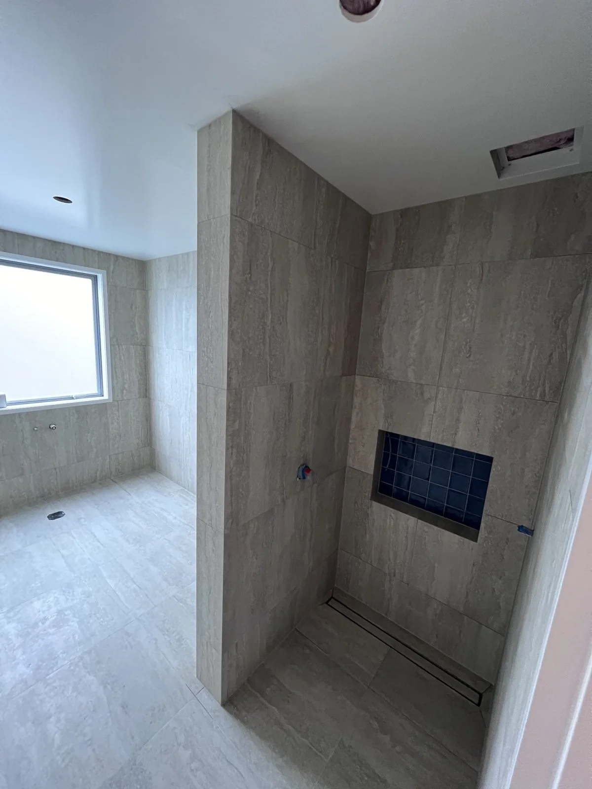 Bathroom with beige tiled walls and floor, featuring a window, a recessed shower area, and a ventilation fan on the ceiling.
