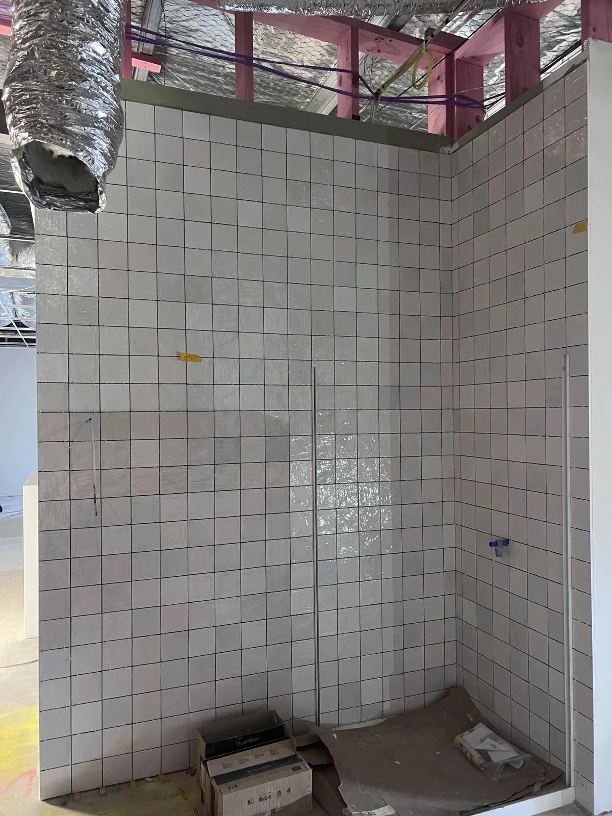 Construction site with tiled wall, exposed ceiling and duct, with tools and materials on the floor.