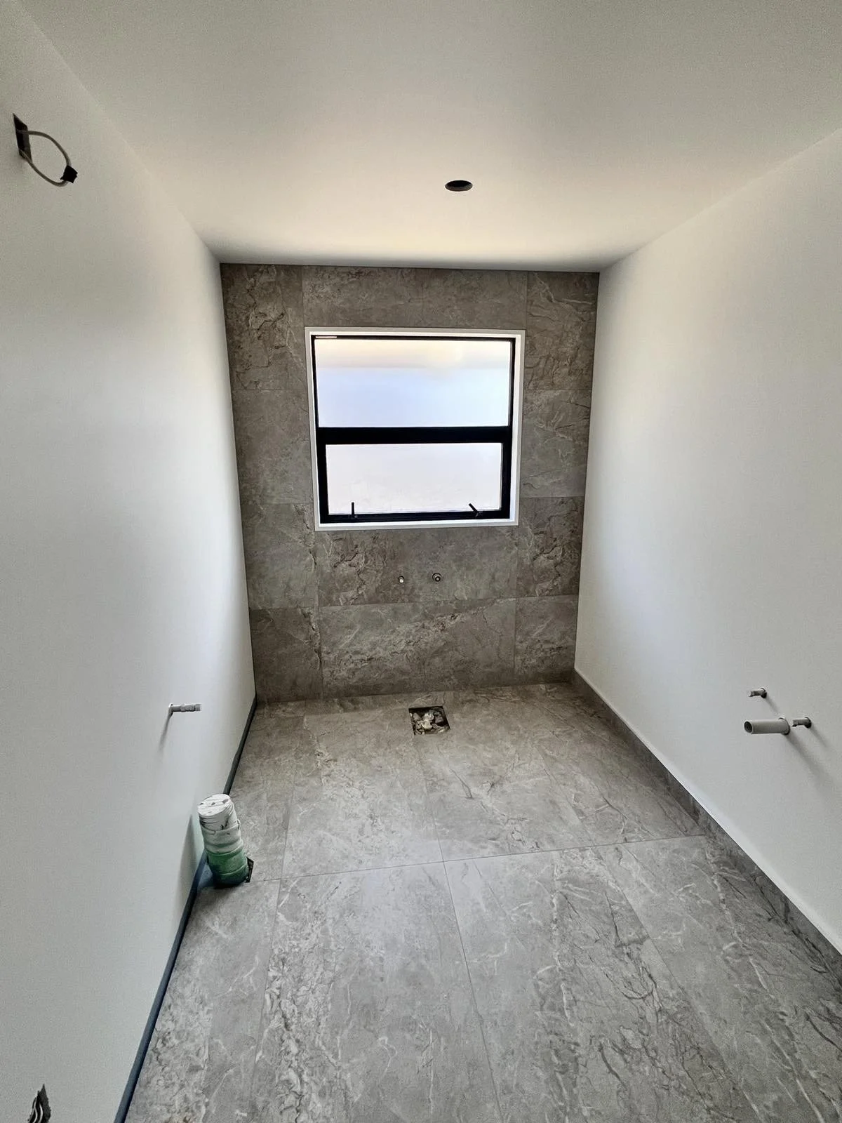 Empty bathroom under construction with tiled floor and walls, a window, no fixtures installed yet, pipes visible on the walls, and construction materials on the floor.