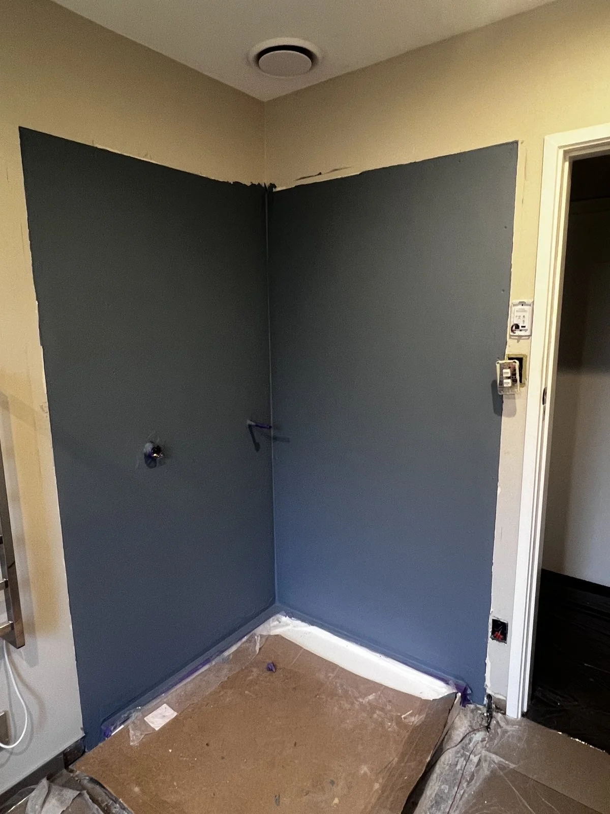 A wall painted dark gray in a room under renovation. The paint job covers two walls at the corner, with edges taped off. Some electrical outlets and switches are visible, and the floor is covered with protective paper.