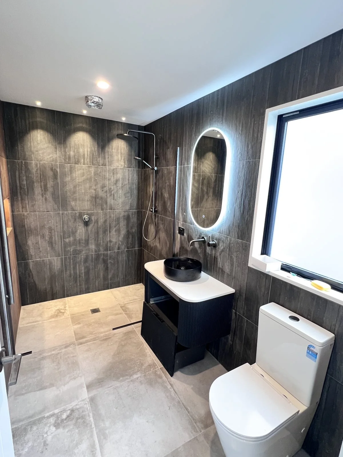 Modern bathroom with dark tiled walls, a walk-in shower, a black vessel sink on a white vanity, an oval backlit mirror, a large window, and a white toilet.
