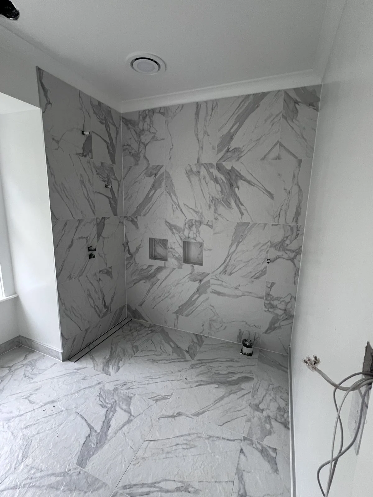 Bathroom with marble tiles on walls and floor, showing a shower area with built-in niche and plumbing fixtures, under construction.