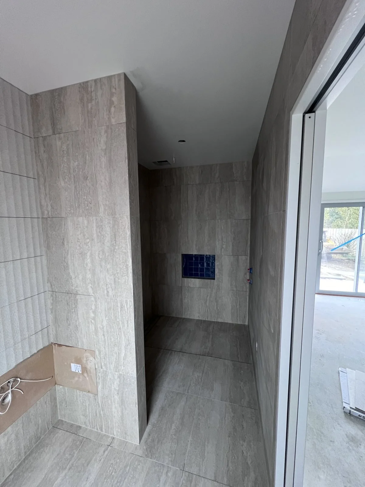An unfinished indoor space with beige tiled walls and floor, a small window with blue tiles, and an open sliding glass door leading outside.