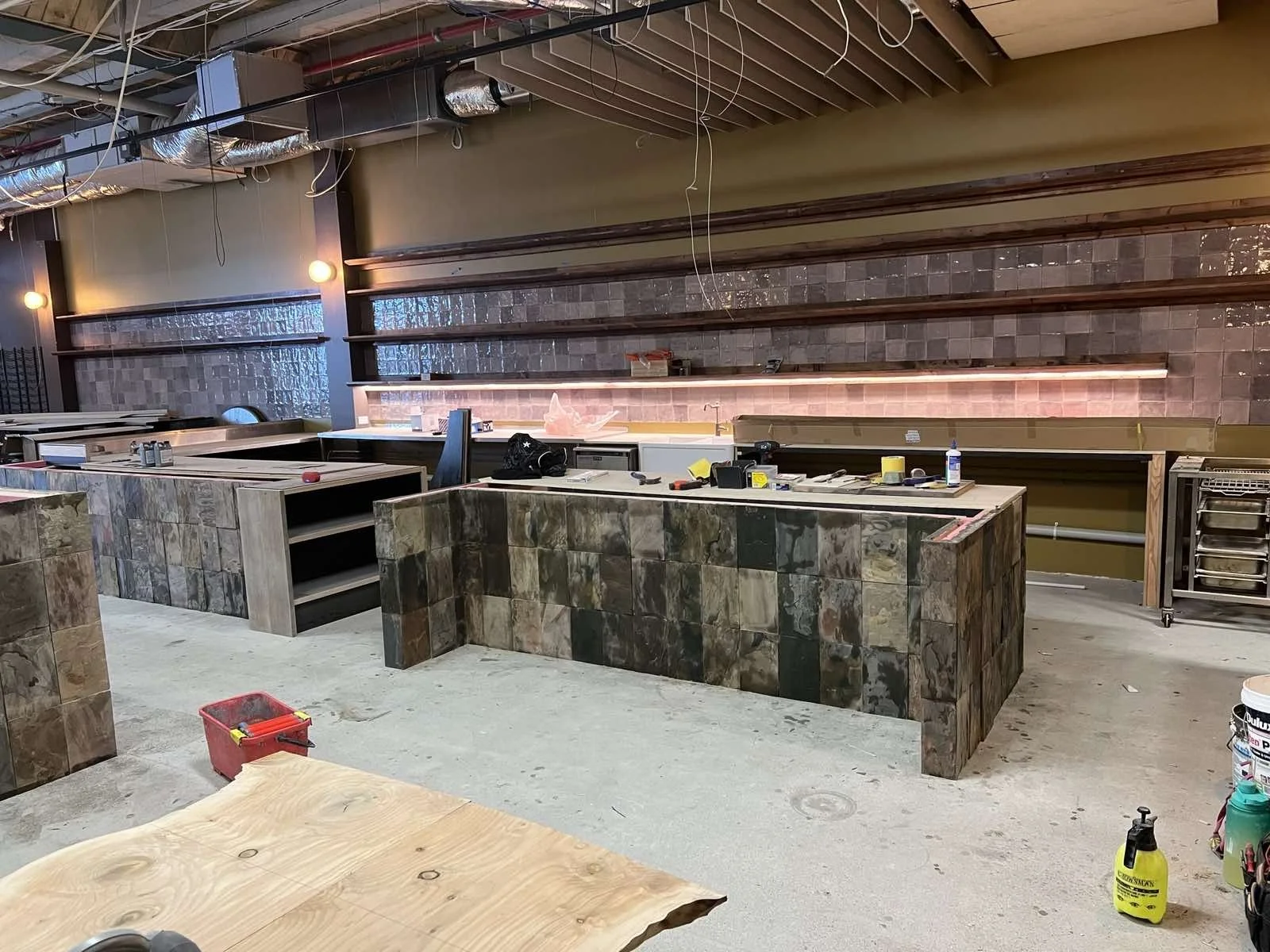 Construction site of a restaurant or cafe interior with partially built island counters and exposed ceiling framework. There are tools, materials, and construction supplies scattered around.