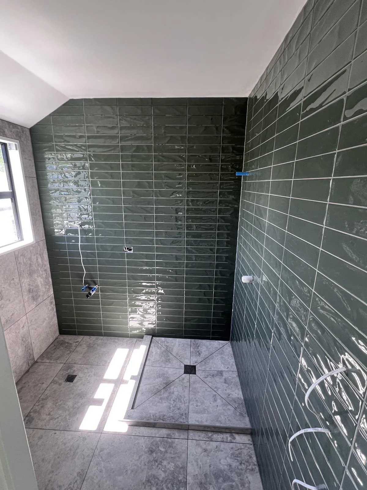An unfinished bathroom with green tiled walls, gray tile flooring, and a window letting in natural light. Plumbing fixtures are in place but no sink or shower installed yet.