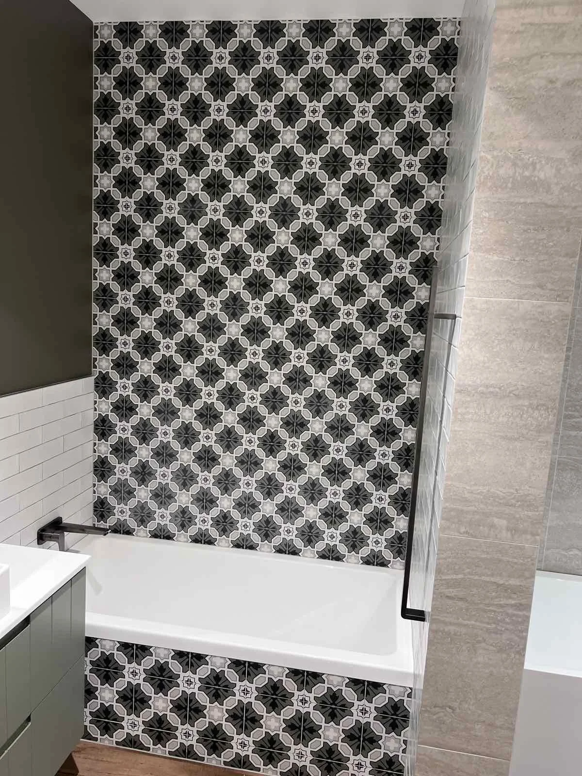 Bathroom with black, white, and gray geometric patterned tiles on the wall and lower section, tan tile on the adjacent wall, and a white bathtub with a black fixture.