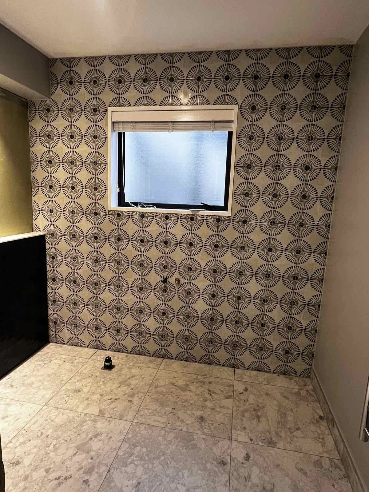Interior room with a patterned wallpaper featuring circular designs, a small window with a closed blind, and beige marble floor tiles.