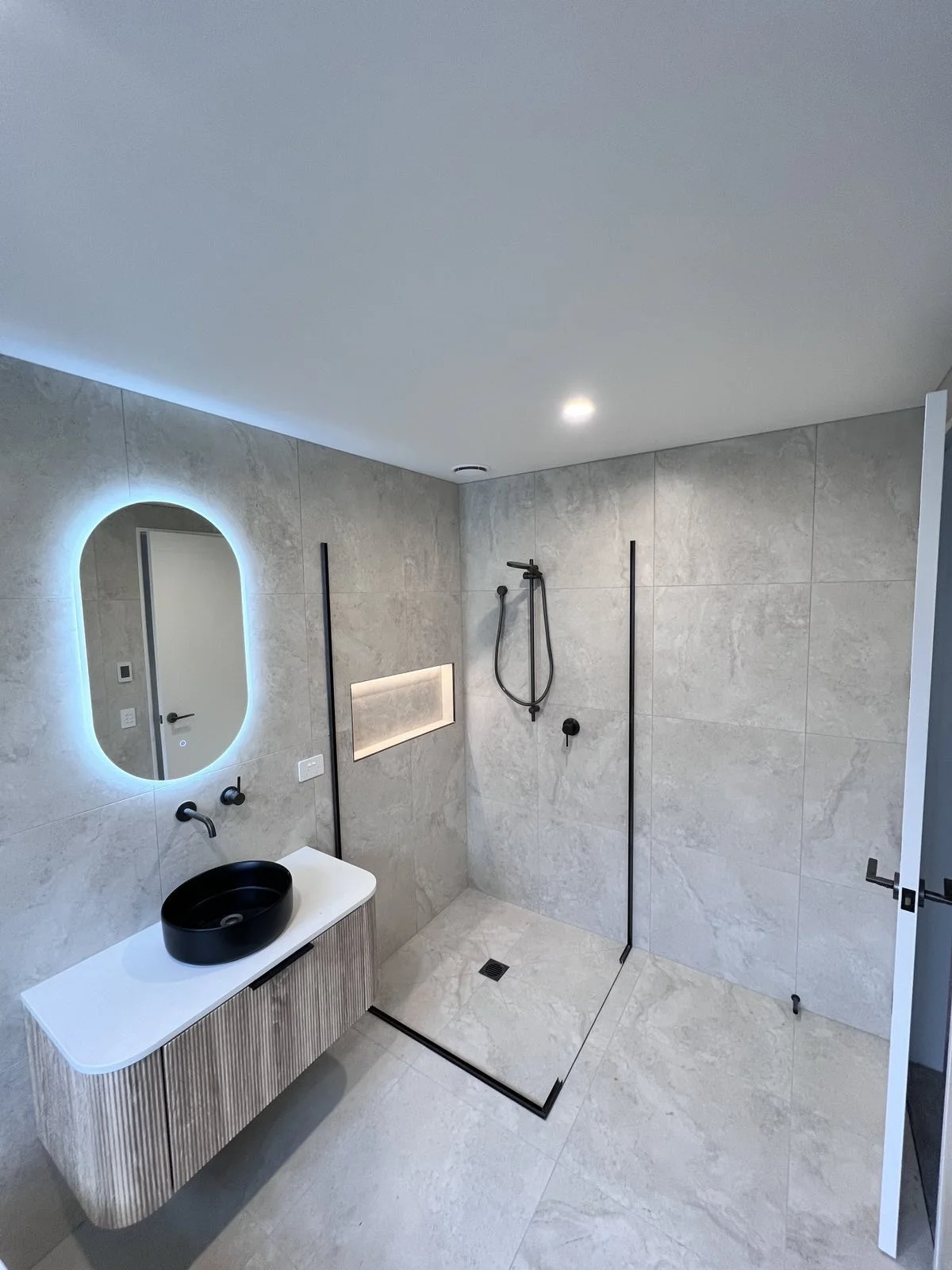 Modern bathroom with light gray tiled walls and floor, black oval sink on a white vanity, illuminated oval mirror, black wall-mounted faucet, shower area with black fixtures, a recessed shelf, and a linear drain on the shower floor.
