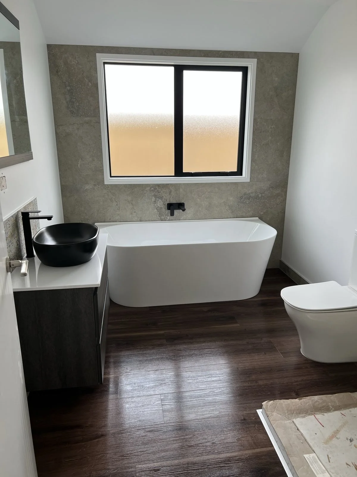 Modern bathroom with a black vessel sink, a white vanity, a clawfoot bathtub under a window, and a toilet on dark wooden flooring.