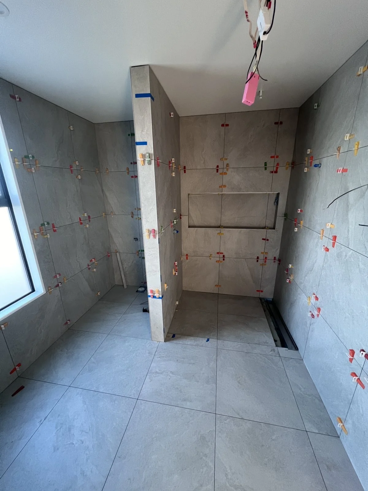 Interior of a room under construction with tiled walls and floor, and colored clips attached to wires for wall tiling, with a window on the left side.