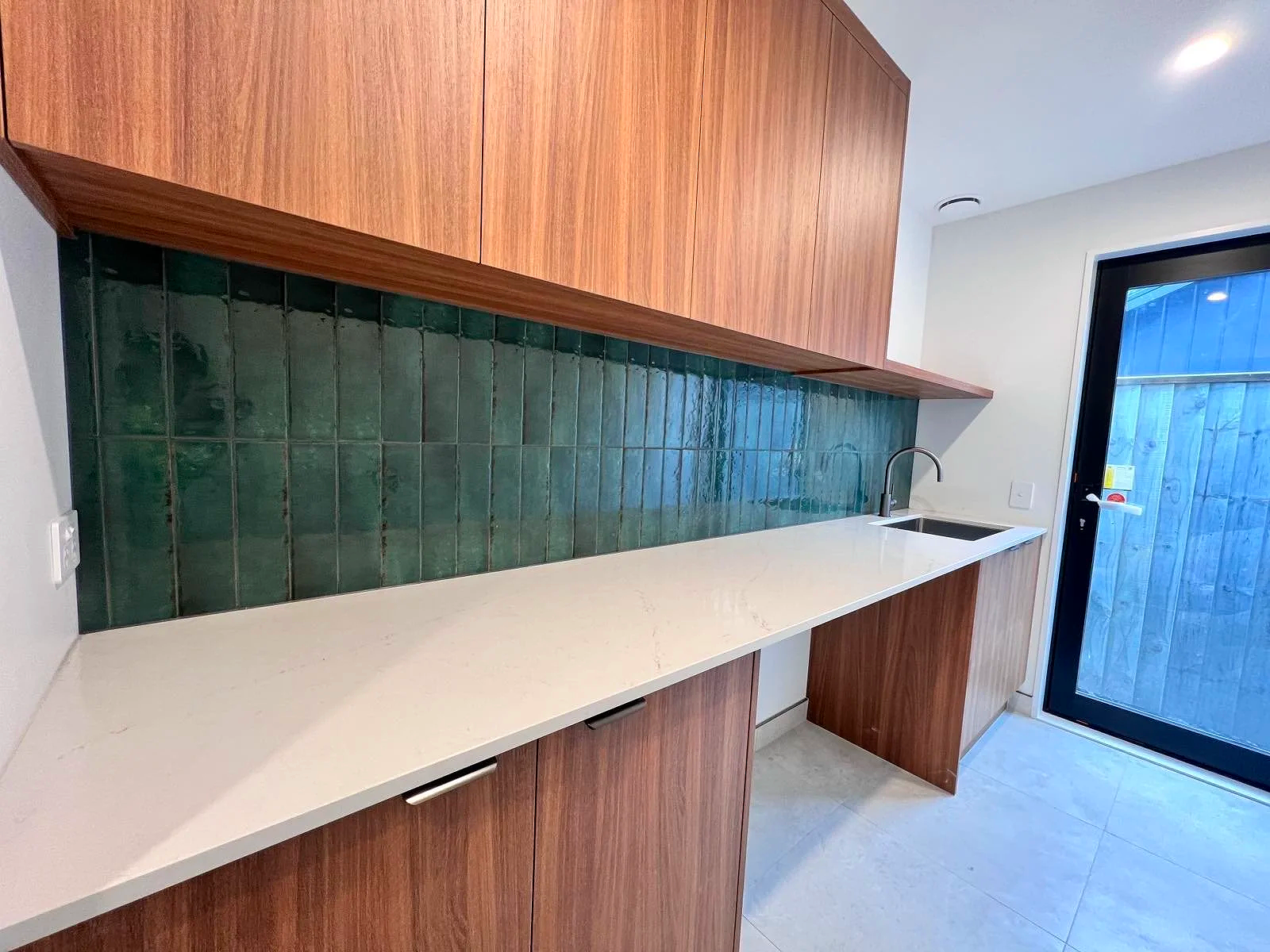 Modern kitchen with white countertop, green tiled backsplash, wooden cabinets, and a black sink. A glass door leads outside.