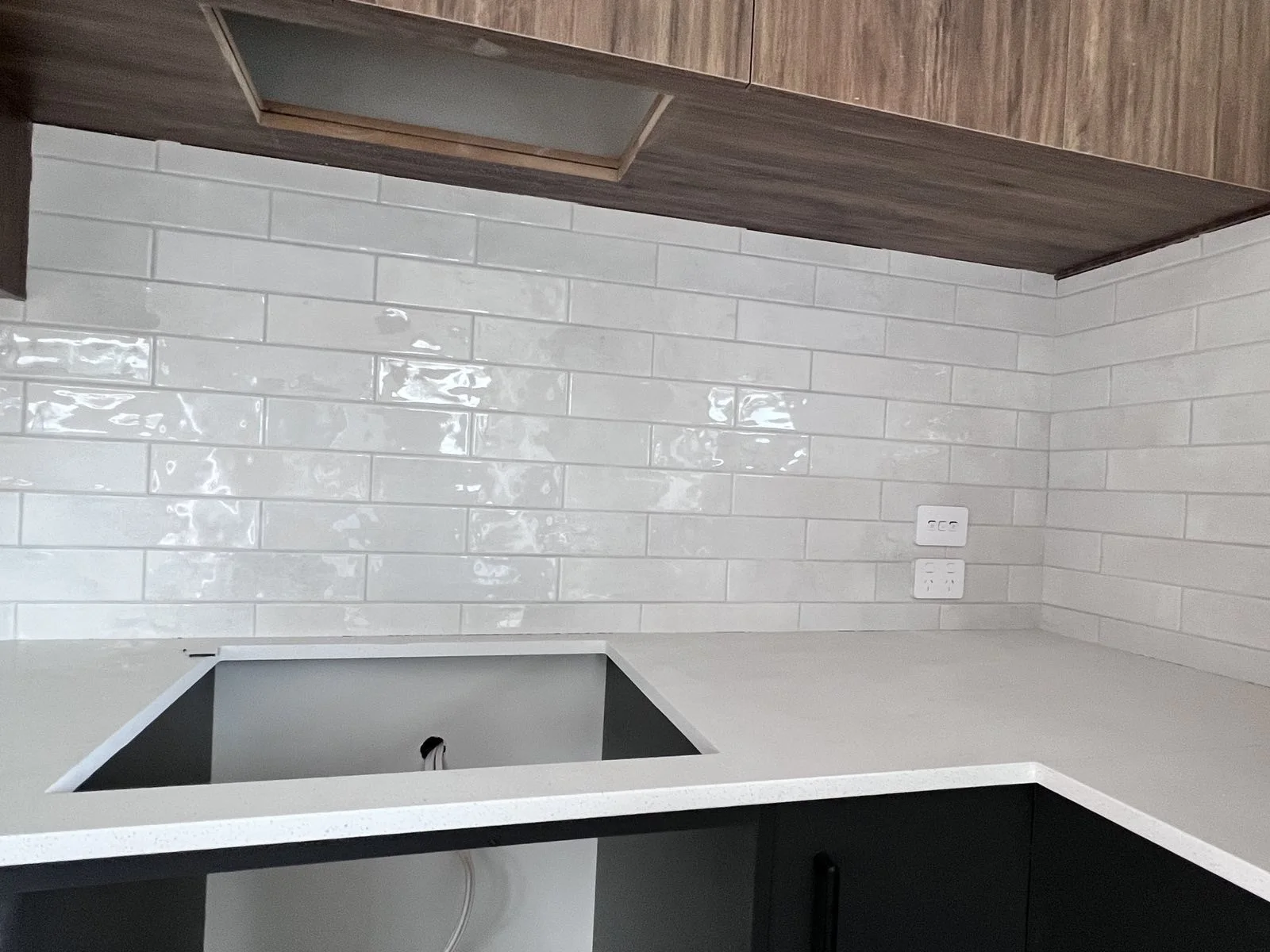 Close-up of a white subway tile backsplash in a kitchen, with a dark cabinet and a partially installed white sink countertop. An electrical outlet and a switch plate are visible on the tiled wall.