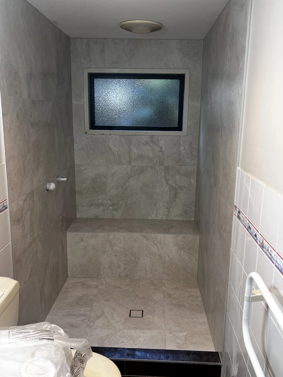 A shower area with beige marble tiles, a frosted glass window, built-in seating, and a white grab bar.