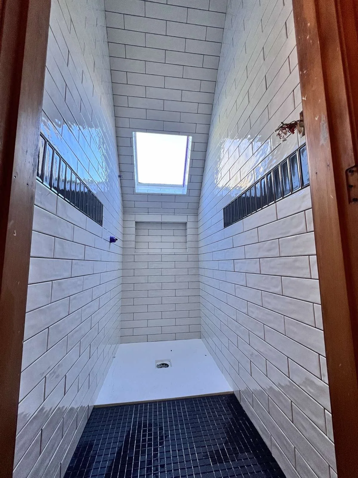Empty shower stall with white brick tiles, black tile floor, a small skylight, and a recessed niche for toiletries.