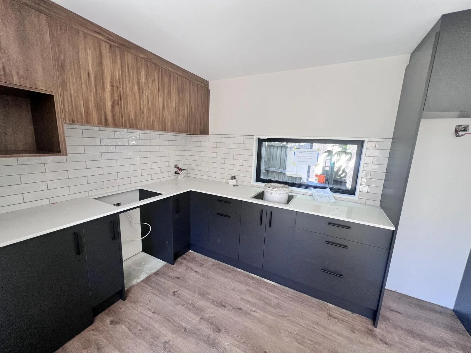 Kitchen under construction with black cabinets, white countertops, white subway tile backsplash, wooden upper cabinets, a window, and construction materials and tools.