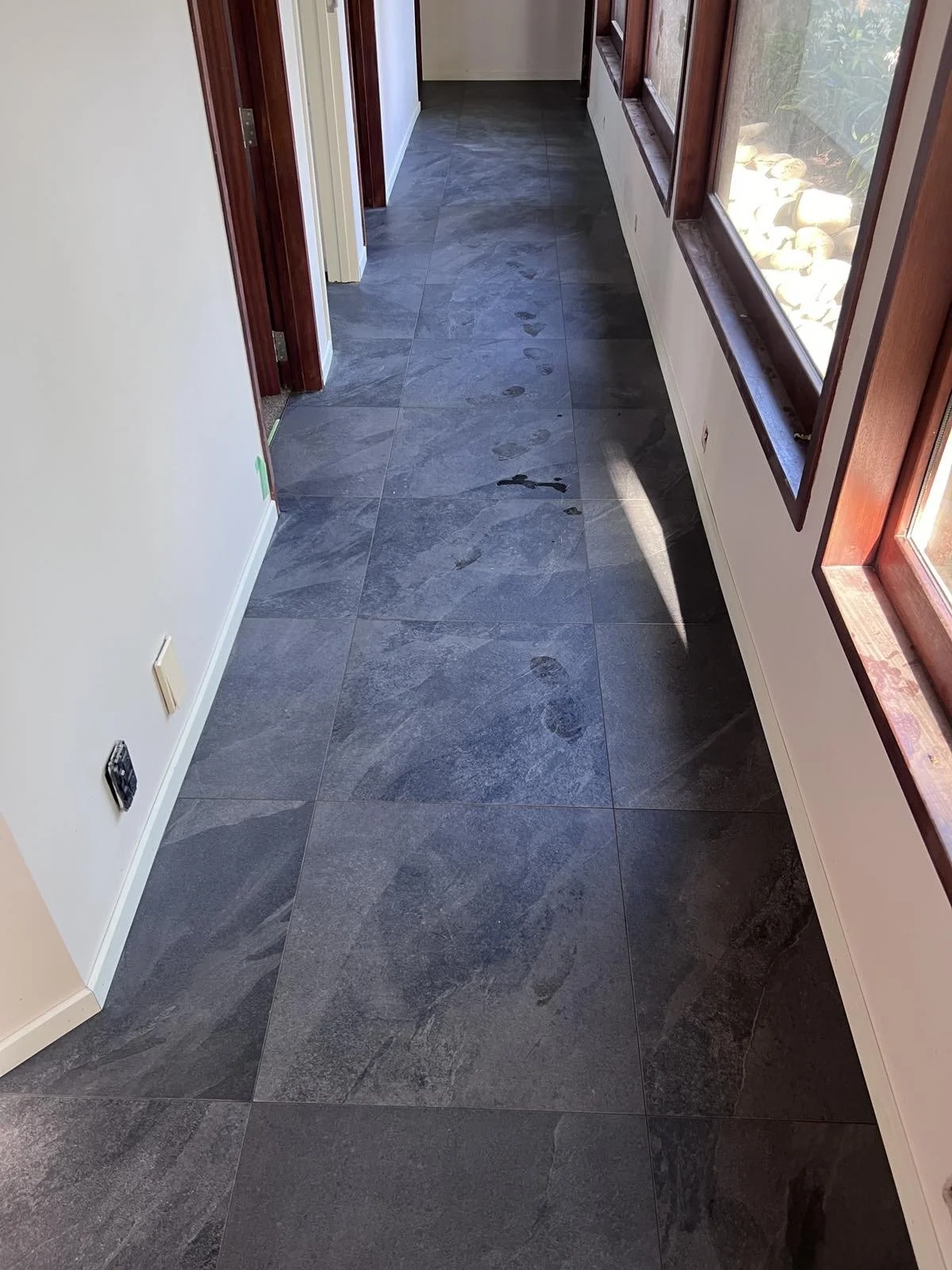 A hallway with dark gray tiled flooring, white walls, and wooden window frames. Footprints are visible on the tiles, and sunlight is shining through the windows.