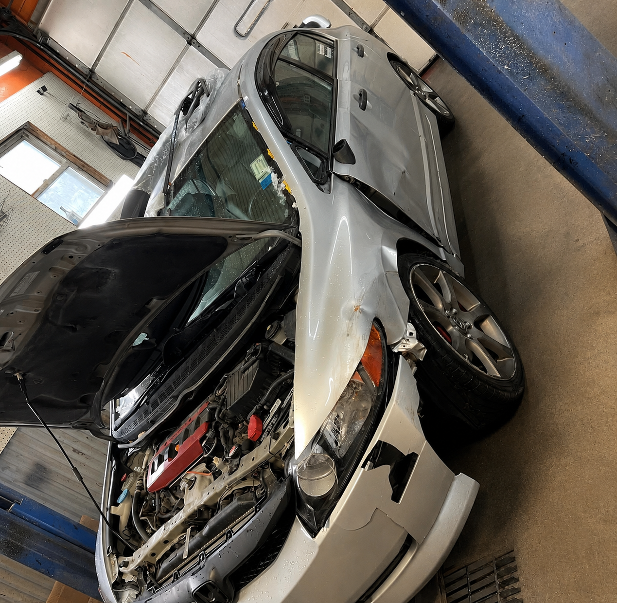Silver sports car with its hood open, showing the engine, in a garage workshop.