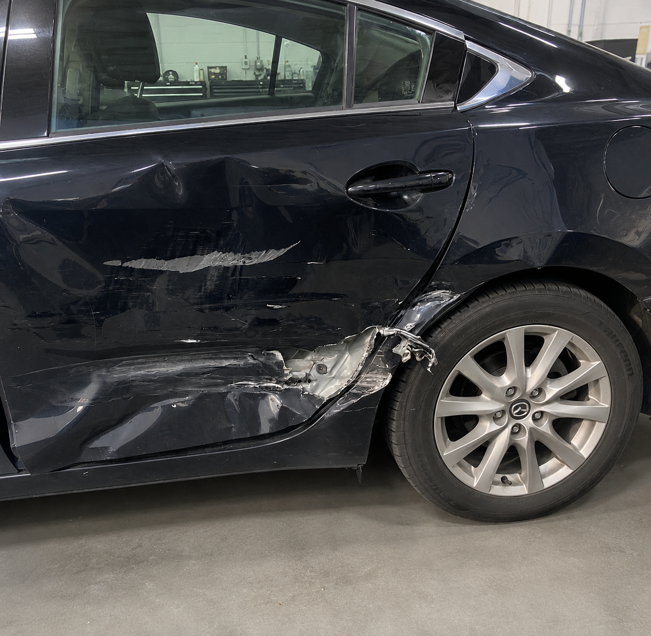 Black sedan car with significant damage to the driver side door and rear quarter panel, showing dents, scratches, and torn metal, parked indoors in a workshop or garage.
