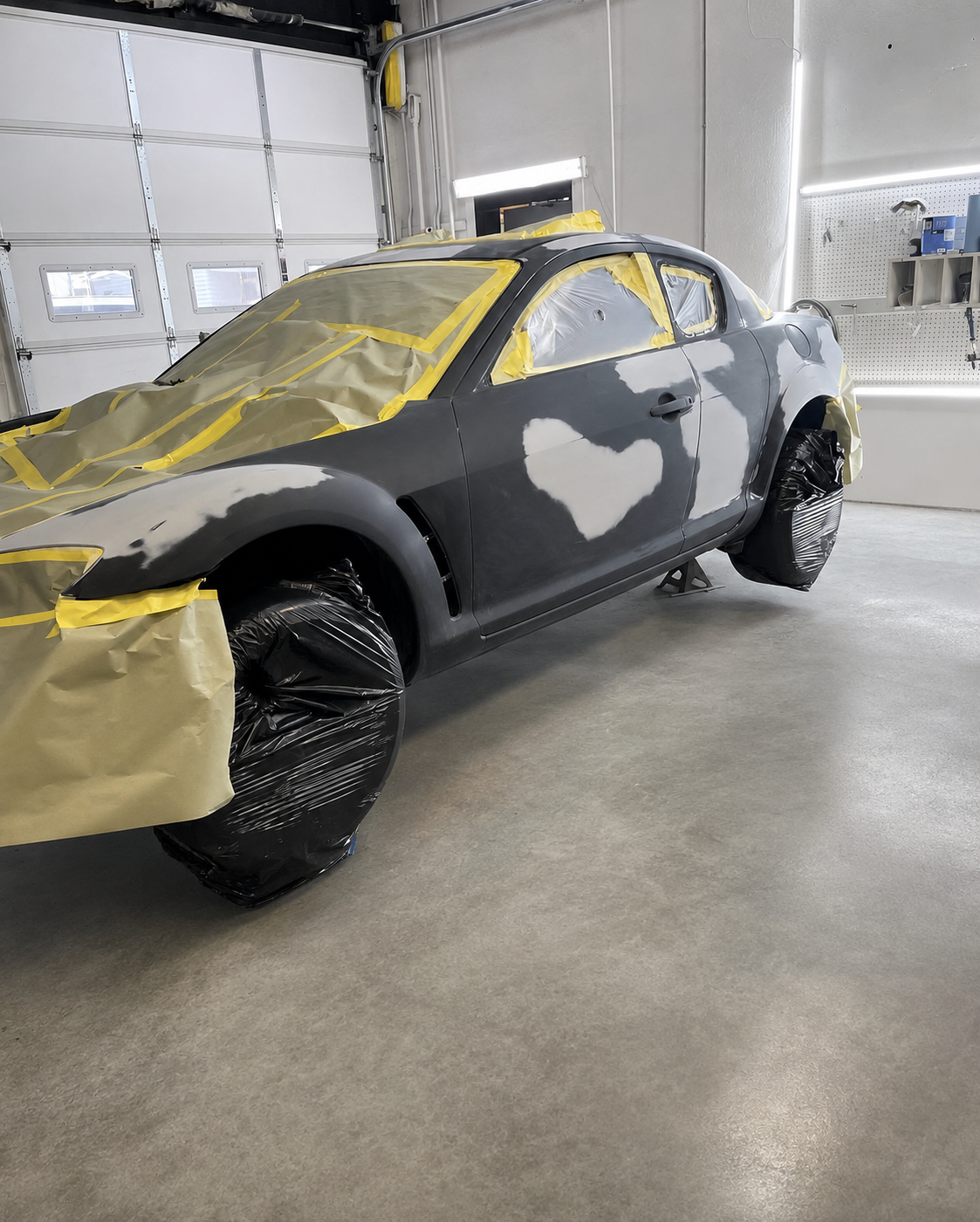 A gray sports car being painted in a garage, with parts covered in masking paper and plastic for painting.