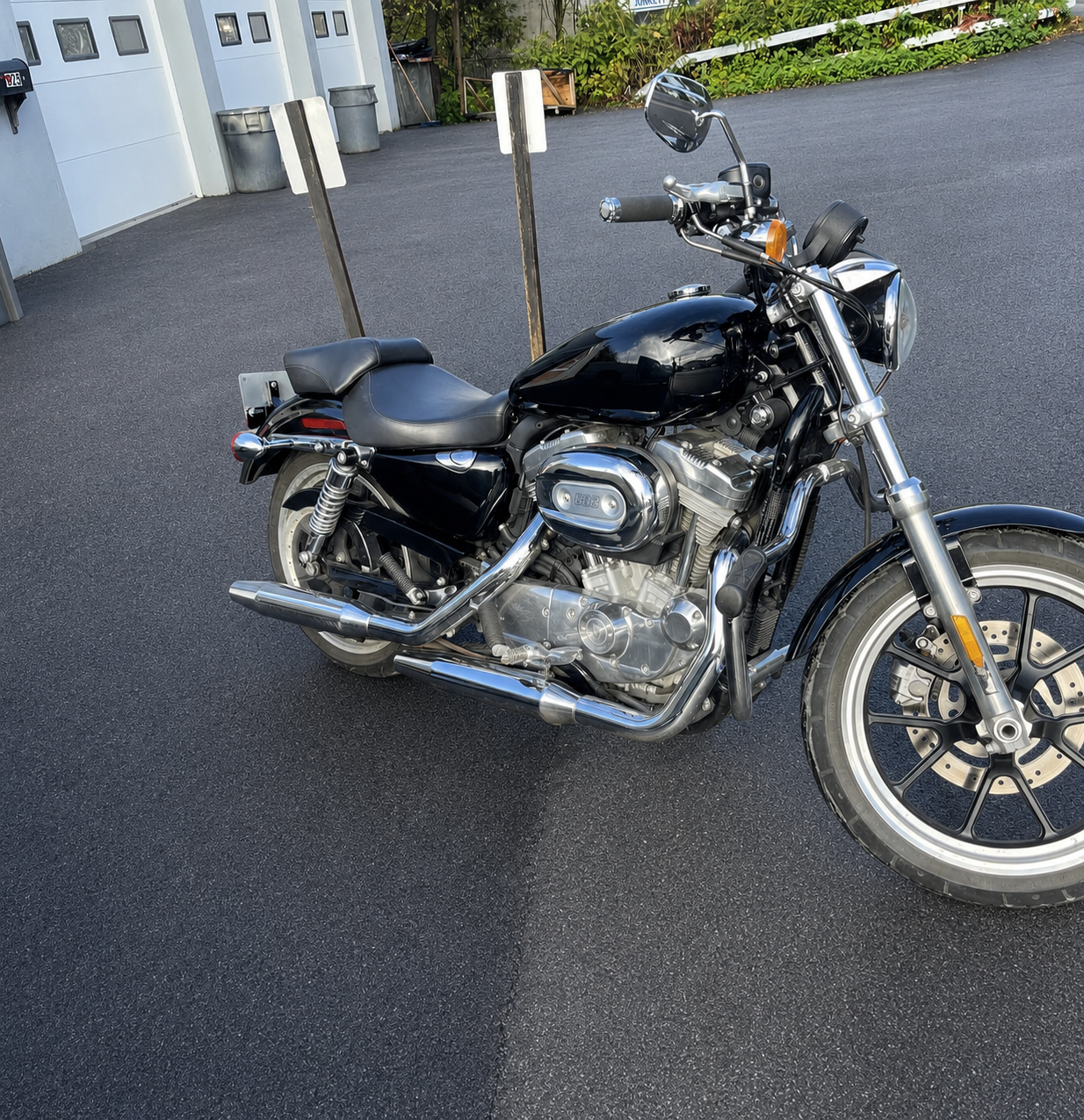 Black and chrome Harley-Davidson motorcycle parked on black asphalt pavement in a lot with garages and trash bins in the background.