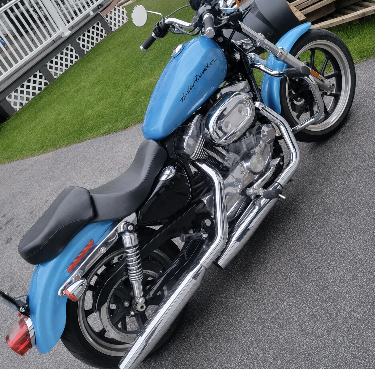 A blue Harley Davidson motorcycle parked on a driveway near a patch of grass and a white fence.