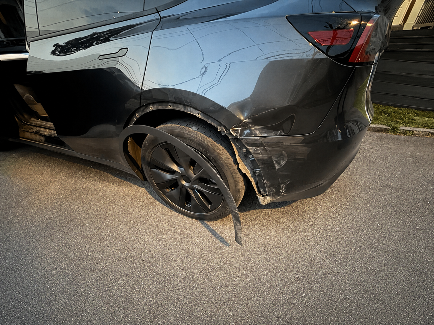 A black car with a damaged front right wheel and fender, showing scratches and a detached fender panel, parked on a street with a black fence in the background.