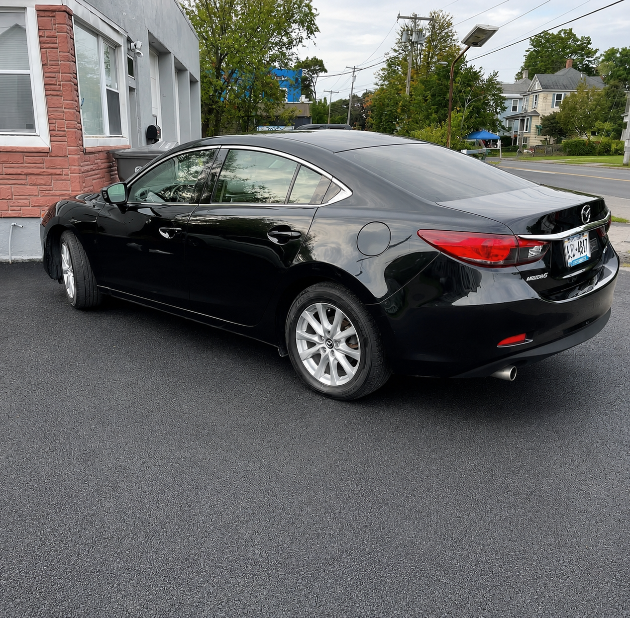 A black Mazda sedan parked on an asphalt lot near a brick building with windows and a trash bin, with residential houses, trees, and power lines in the background.