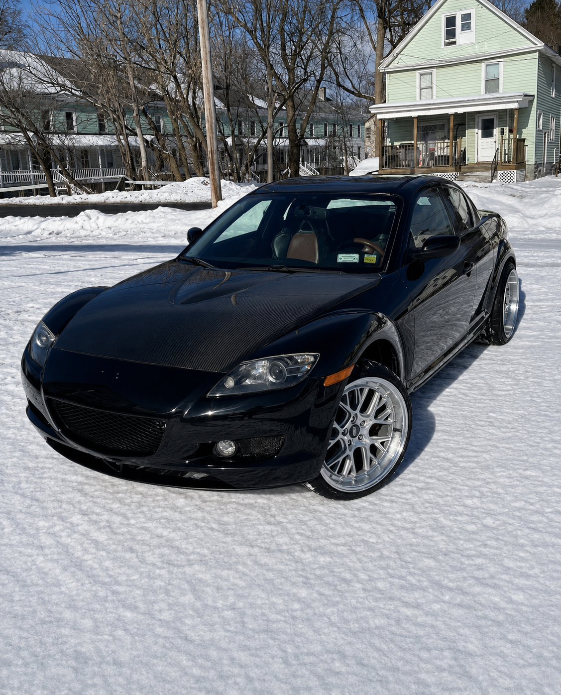 A black sports car parked on snow-covered ground in a residential neighborhood on a sunny winter day.