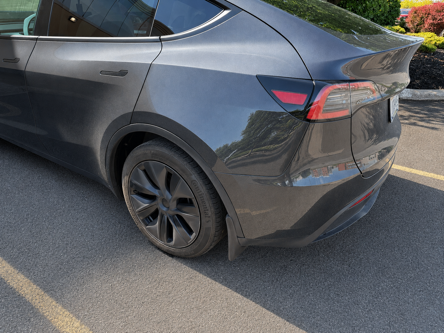 The rear quarter of a dark gray electric car, parked in a parking lot, showing the rear wheel, taillight, and part of the trunk and rear window.